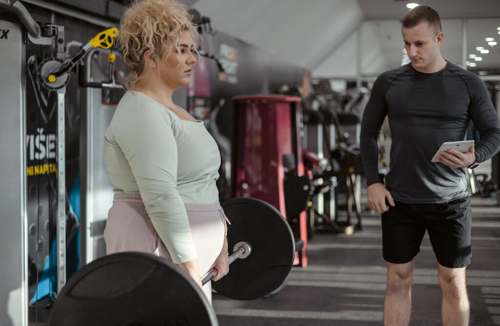 Concept of Weight Loss. Plus Size Woman in Sportswear Doing Weight Exercises With Assistance of Her Personal Trainer at Gym With Modern Fitness Equipment for Fitness