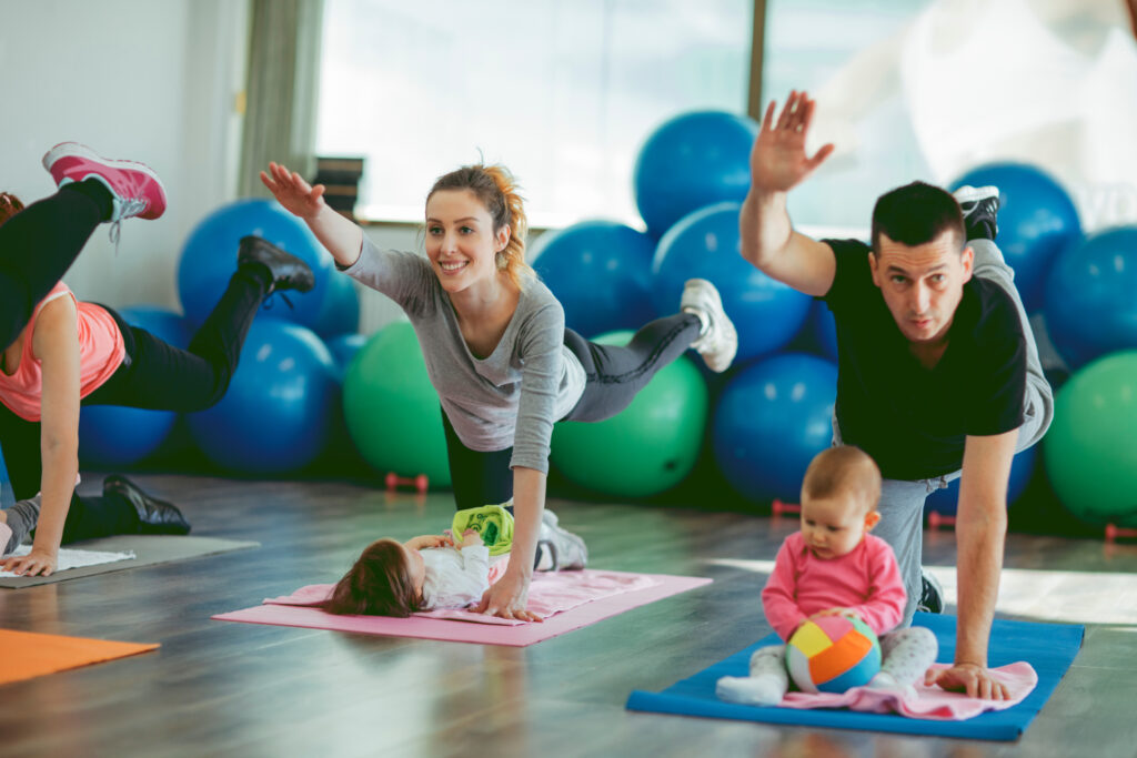 Parents Exercising with Their Babies in a Gym. Selective focus on father exercising with his baby. He is exercising on floor on exercising mat and baby is sitting in front of her dad. He is happy to exercise together with his baby and stay fit.