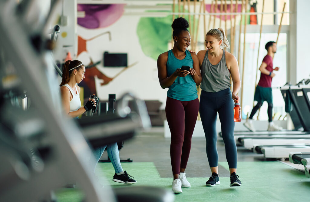 Happy athletic women having fun while using cell phone during sports training in a gym.